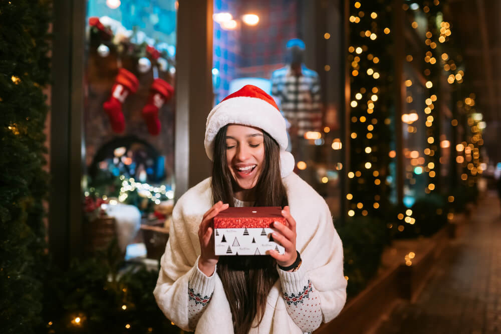 A girl wearing a Santa hat is laughing with a Christmas gift box on the street